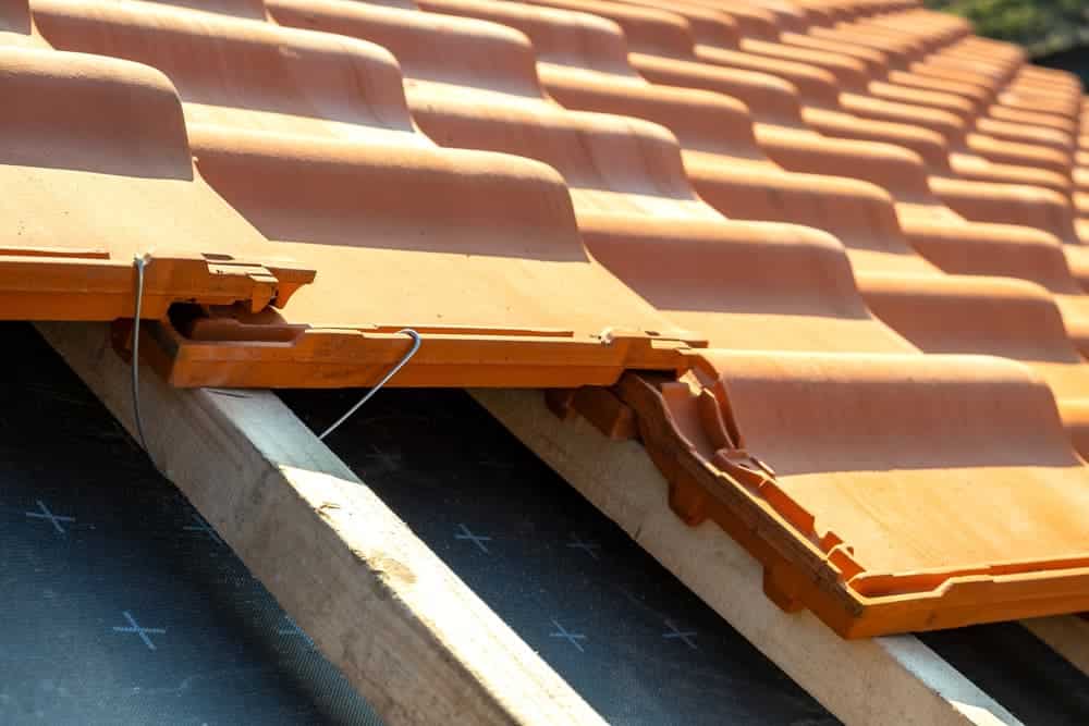 Closeup of metal montage anchor for installation of yellow ceramic roofing tiles mounted on wooden boards covering residential building roof under construction.